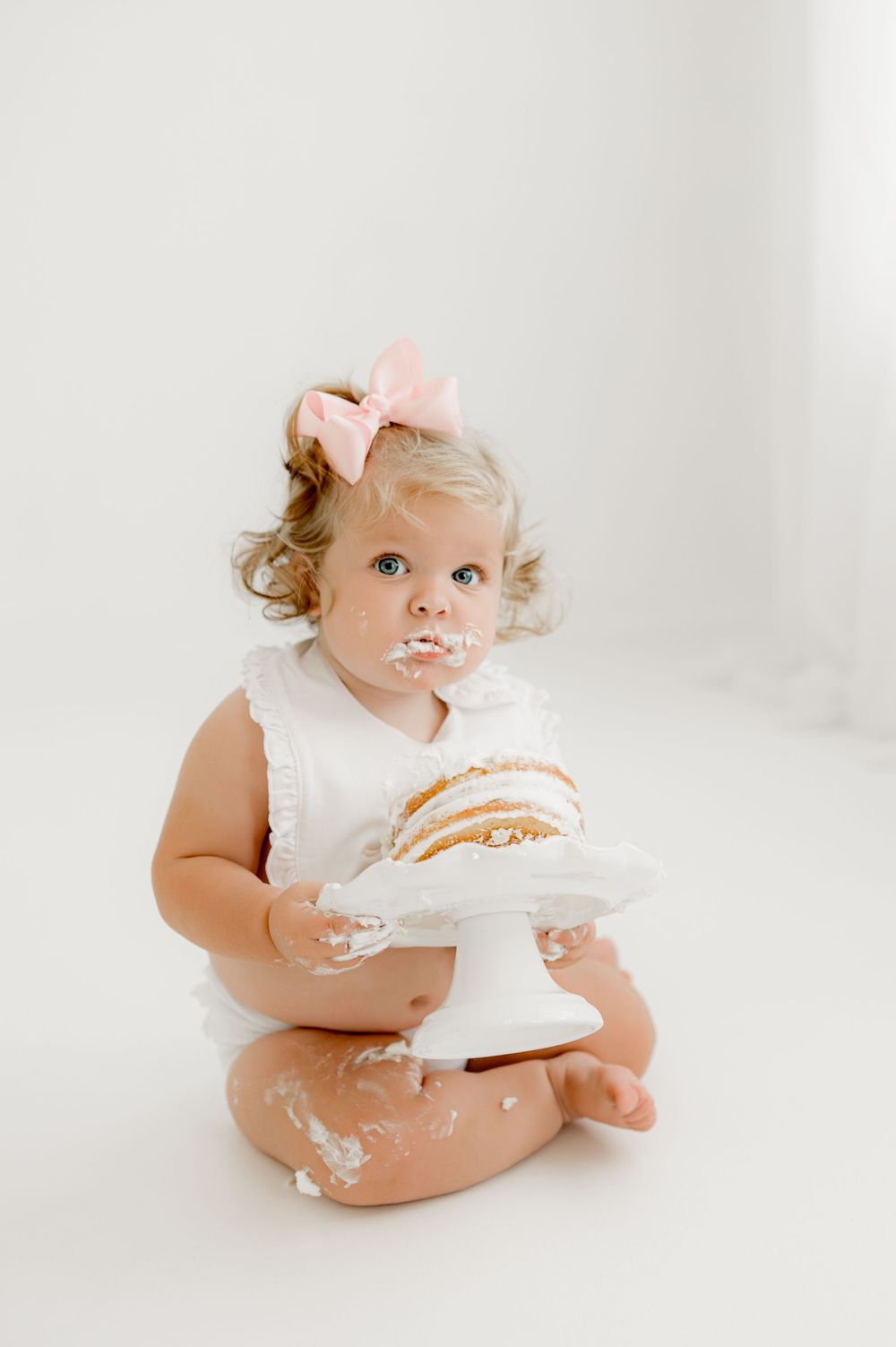 A toddler in a white sleeveless dress sits on white floor with a cake stand, covered in cake crumbs and frosting.