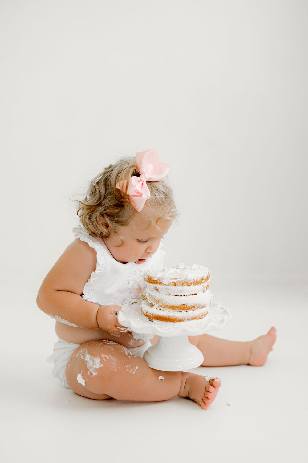Baby in white dress sits on floor with decorated white cake on stand, getting messy during cake smash photo session.