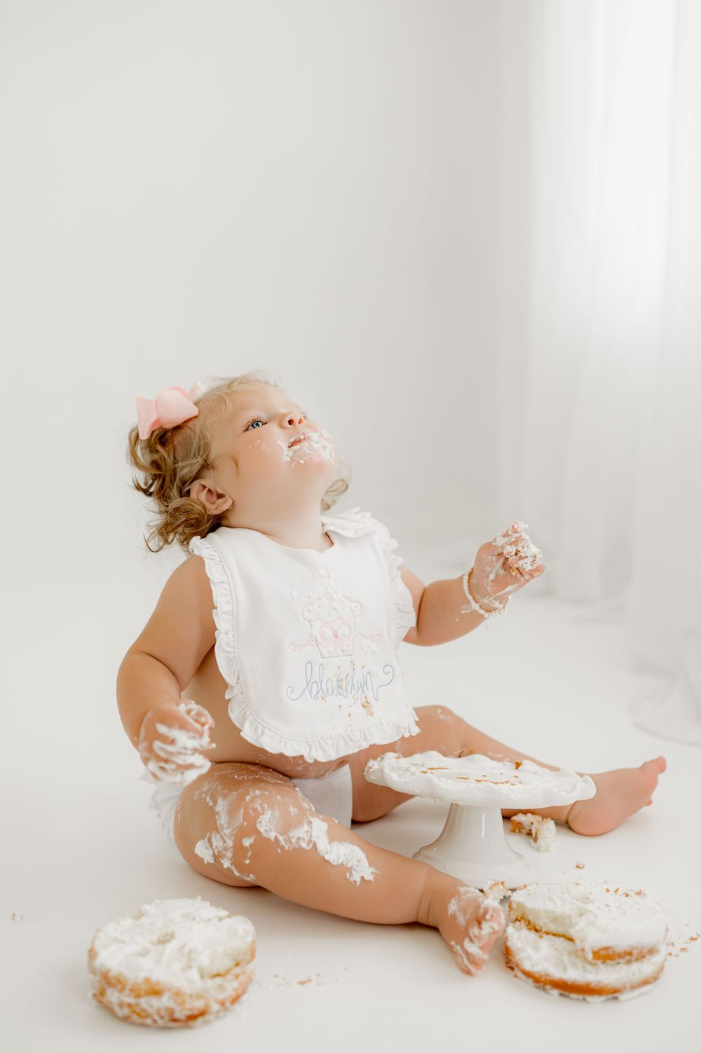 Baby wearing a white bib sits on floor with powdered donuts, playfully covered in powdered sugar during messy snacktime.