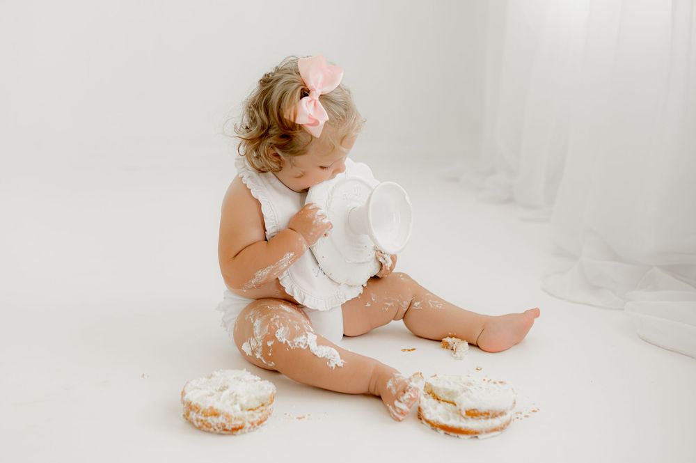 A toddler sits on a white floor with cream-filled pastries and feathers scattered around creating a messy, playful scene.