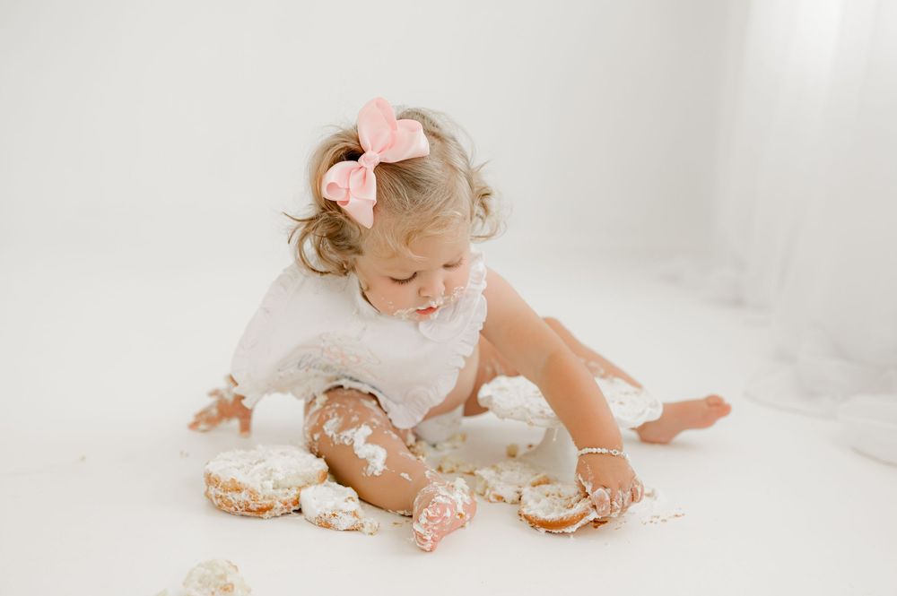 Small child in white dress plays in spilled flour and donuts during minimalist cake smash photo session.
