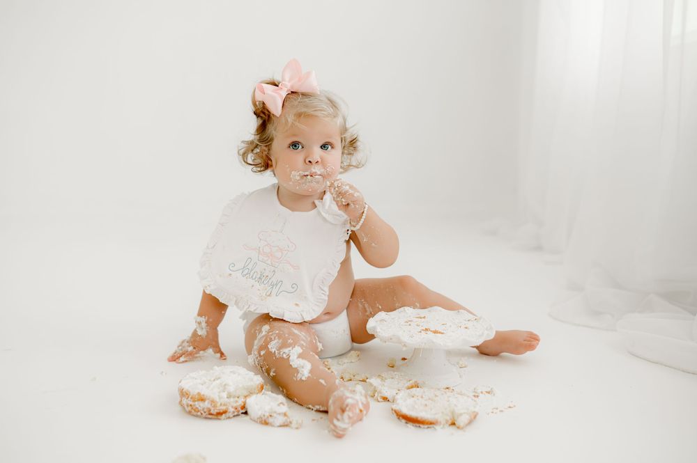 Little one in white outfit sits on floor surrounded by scattered white donuts and snacks on light background.
