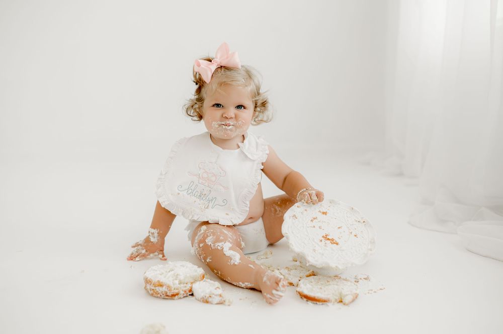 Baby in white dress sitting and playing with birthday cake in bright photography studio setting.