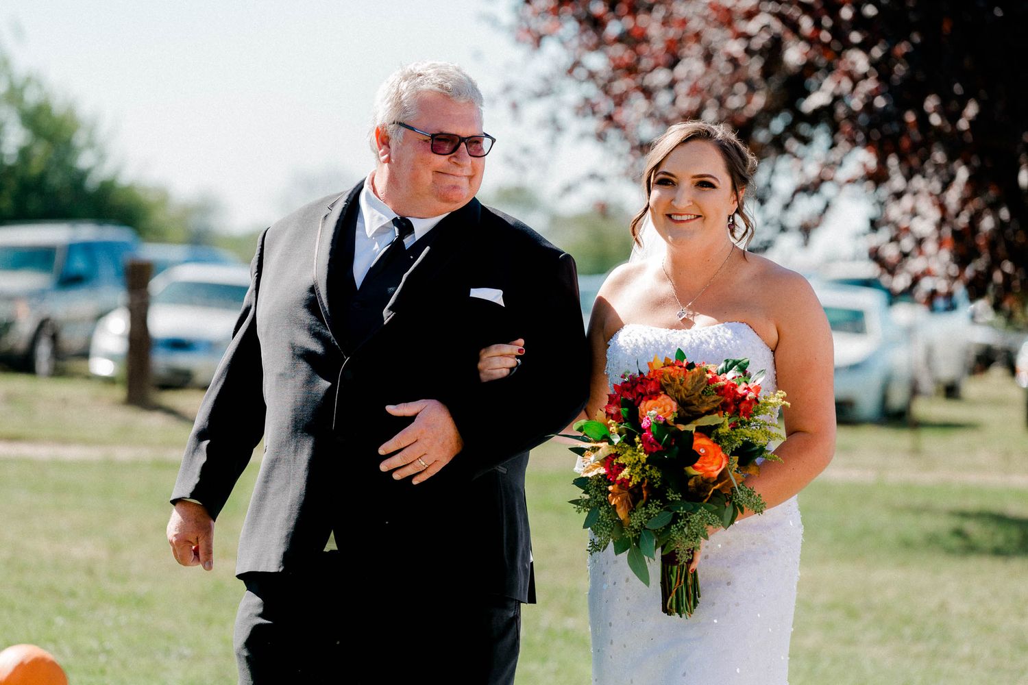 A bride and her dad walks arm in arm down the aisle, during a wedding ceremony, with the bride holding an orange and red bouquet.