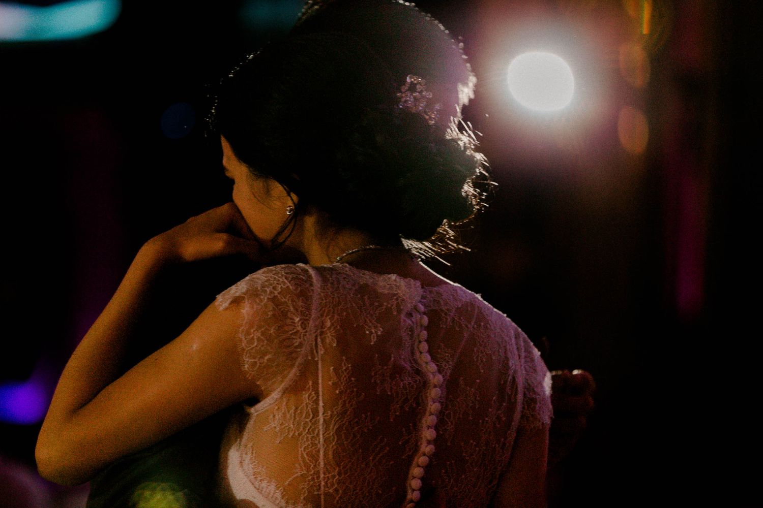 The bride in a lace dress is having the first dance with the groom, illuminated from behind with a blurred bokeh light in the background.