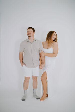 Couple in casual white outfits posing against plain backdrop in minimalist studio setting.