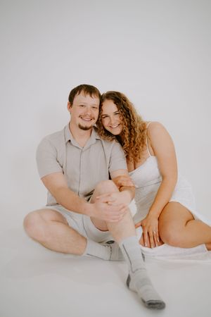 Casual couple sitting together on white floor in studio showing genuine affection.