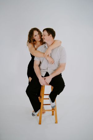 A romantic couple embraces on a wooden stool against a white backdrop during an intimate photoshoot.