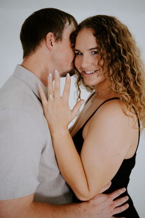 A couple shares an intimate moment while showing off an engagement ring against a white backdrop.