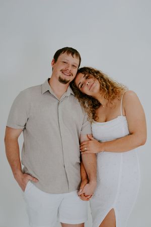 Couple in light colored clothing posing playfully against white studio background.