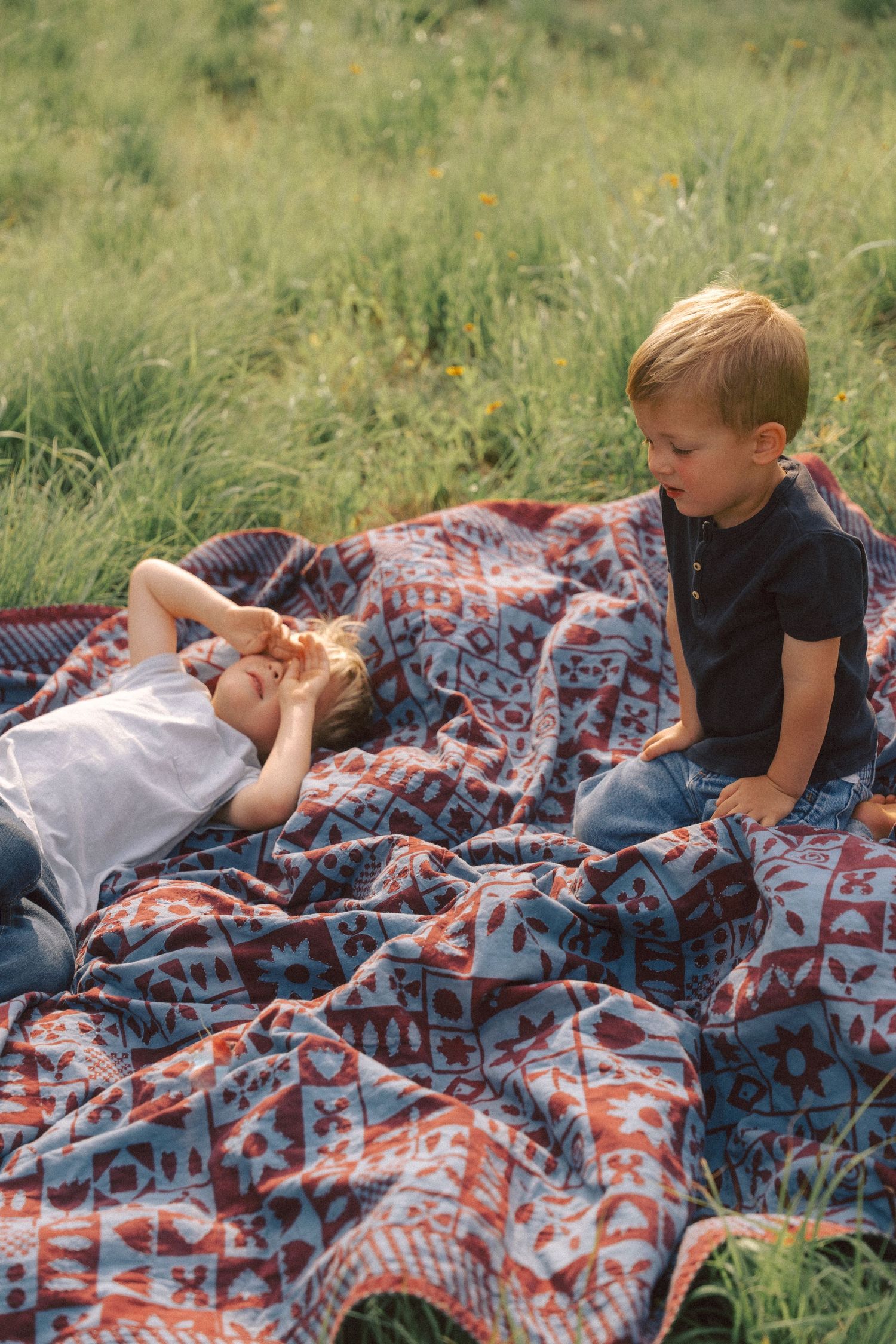 Two people relaxing on a colorful patterned blanket in a grassy meadow on a sunny day.