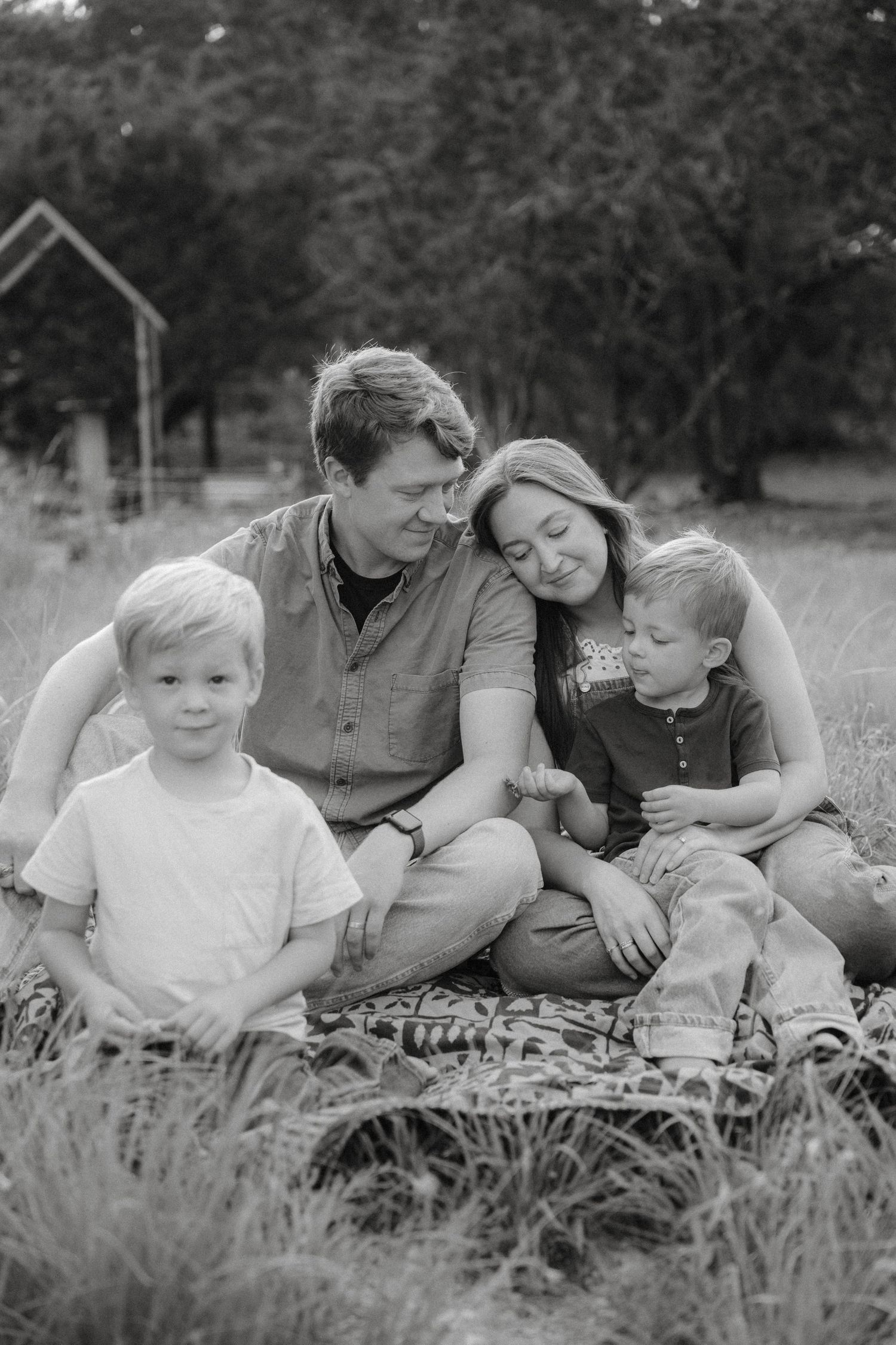 A family sits together on a blanket in tall grass during a black and white outdoor photo session.