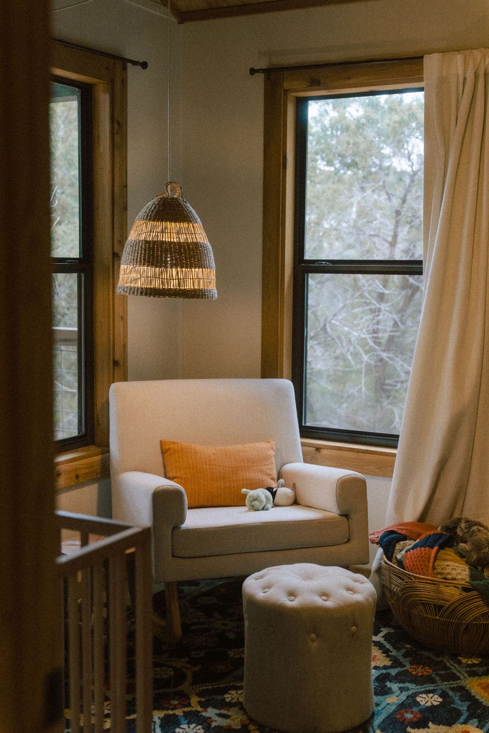 A cozy reading nook with white armchair, woven pendant light, and orange accent pillow by large windows.
