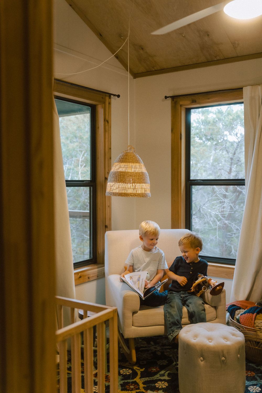 Two children sharing a cozy reading nook with a woven pendant light and bright windows in a modern living space.