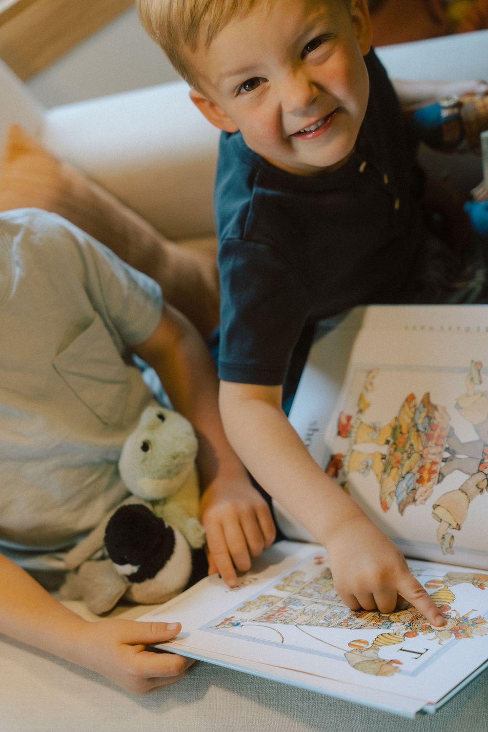 Young child reading a picture book with stuffed animals while lying down in a cozy setting with warm lighting.