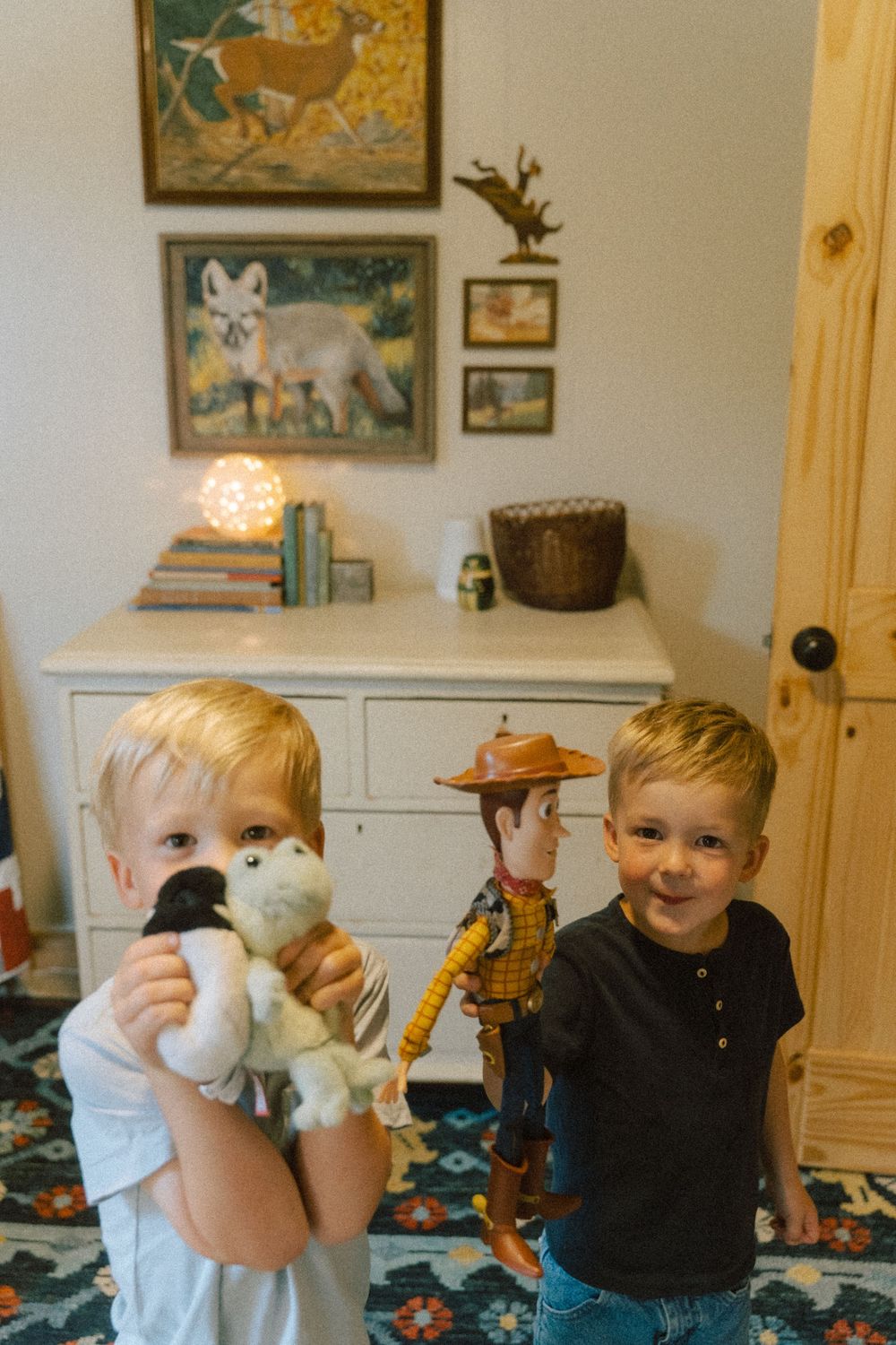 Young children play with soft toys and a Woody doll from Toy Story in a room decorated with artwork and furniture.