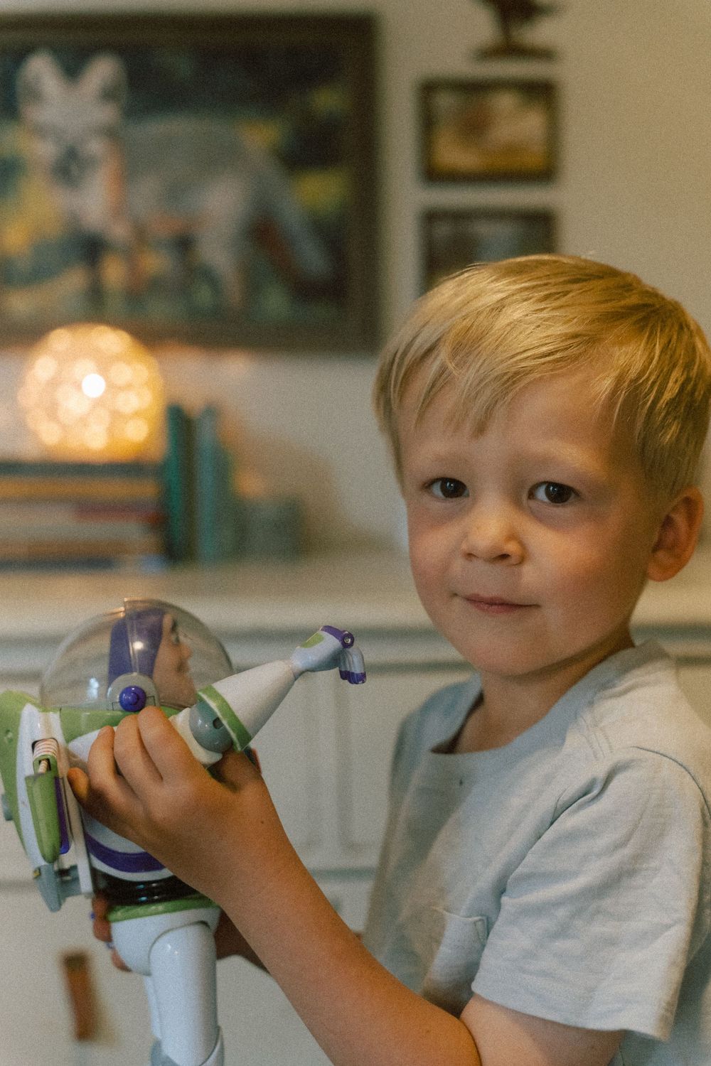 Child holding a Buzz Lightyear toy while sitting indoors with soft lighting and decor in the background.