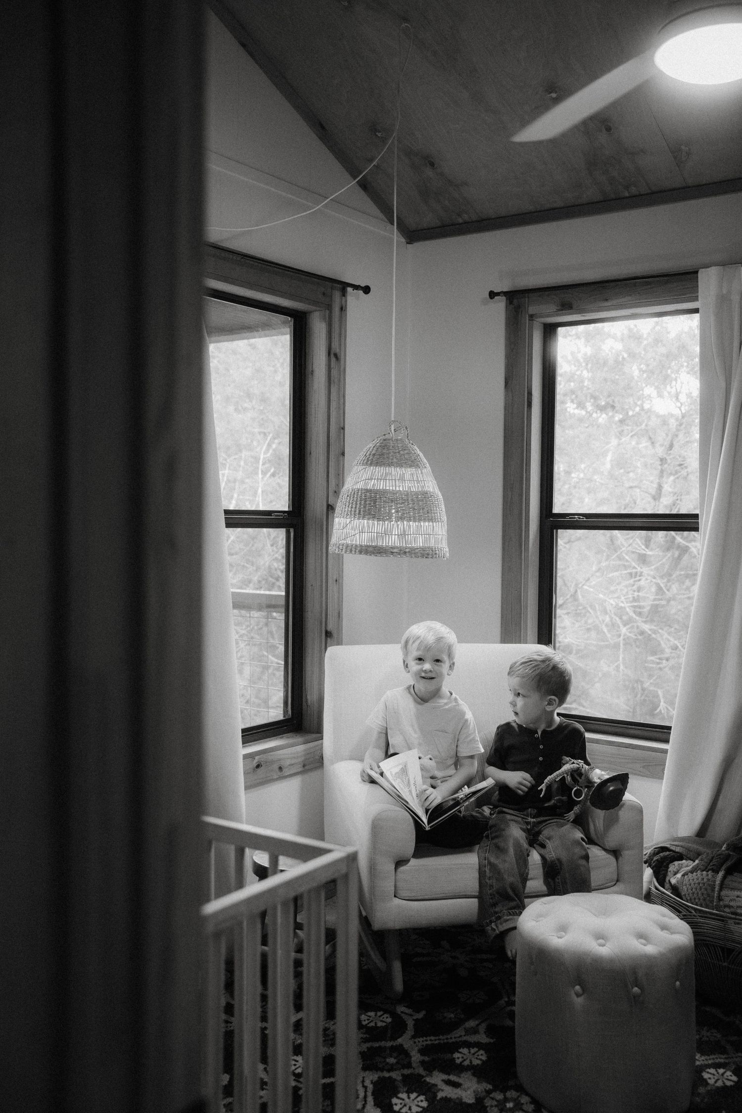 Two small children sit together on a white armchair by windows, with a hanging wicker lamp above in a cozy room.