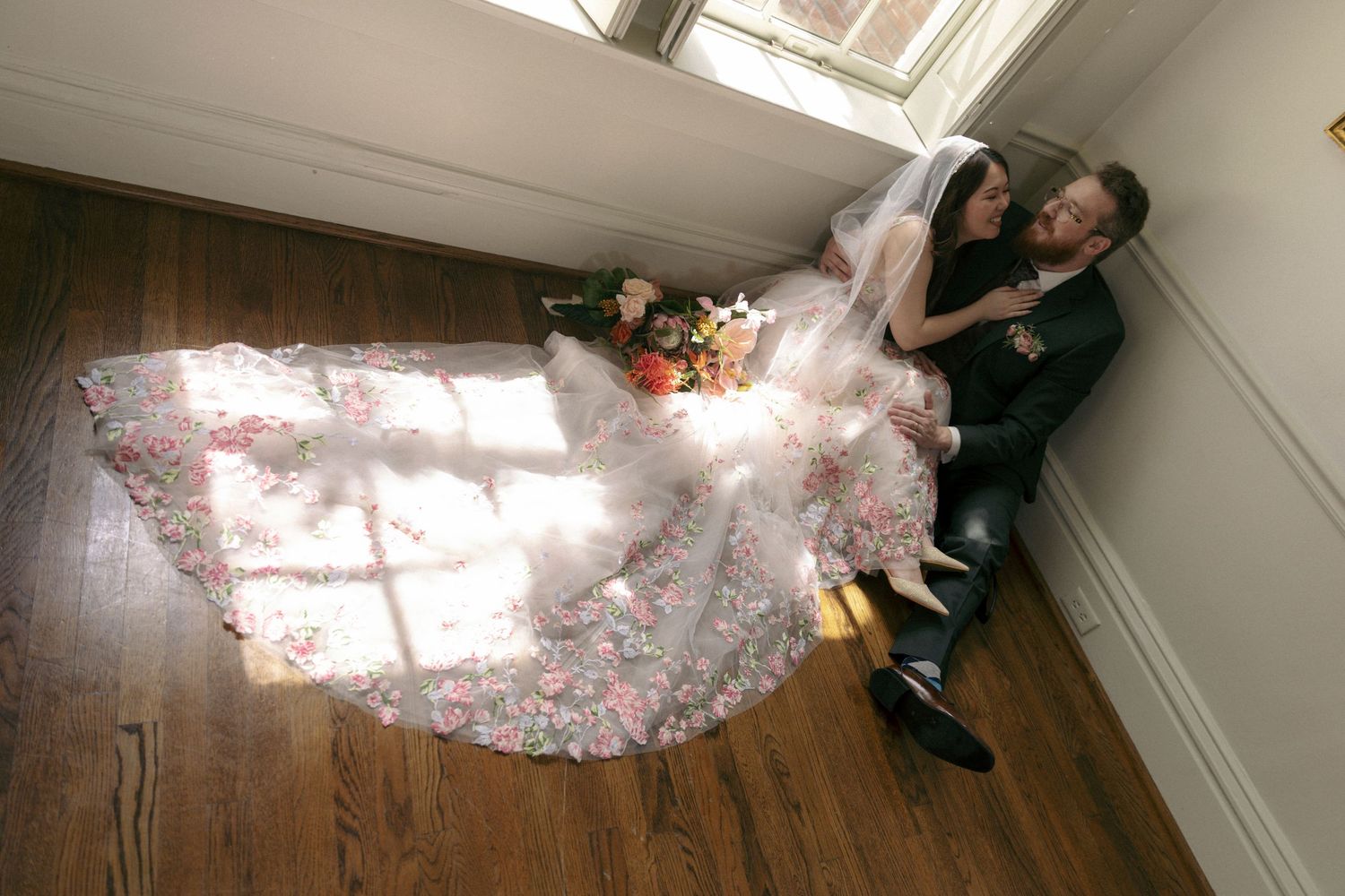 A couple poses on wooden floors in bridal attire as sunlight streams through a window onto a flowing floral dress.