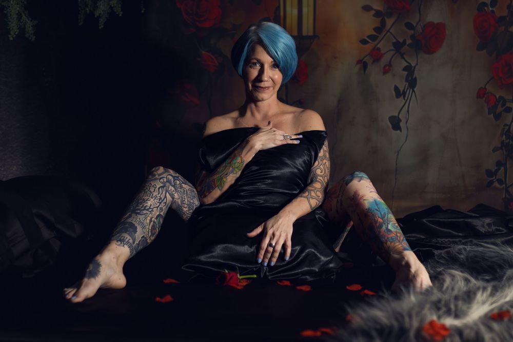 Stylish figure in black dress reclines on dark fabric with floral pattern wall in moody lighting during her empowering boudoir session in TX