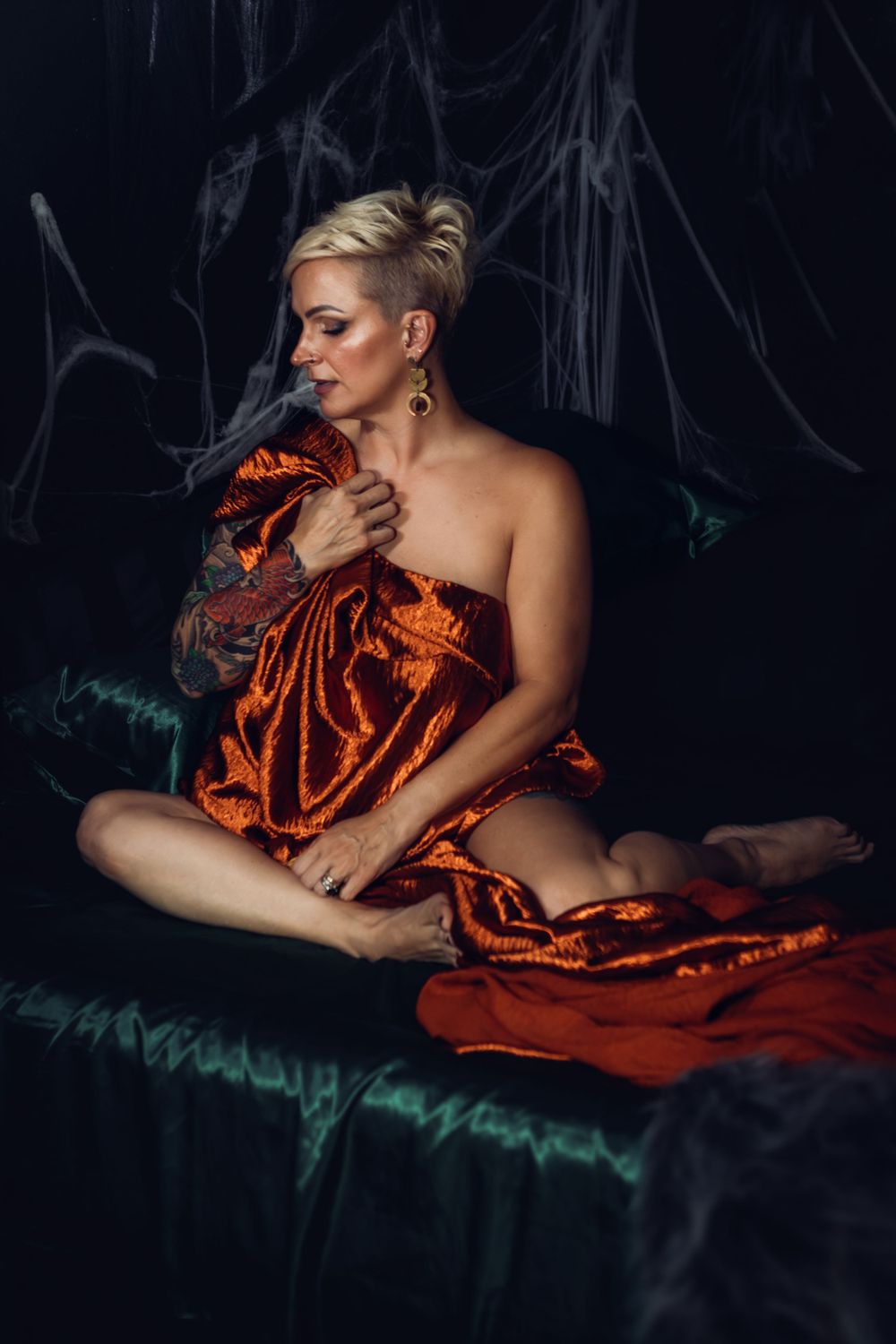 An artistic boudoir portrait featuring someone draped in luxurious burnt orange fabric against a dark background with ethereal elements.