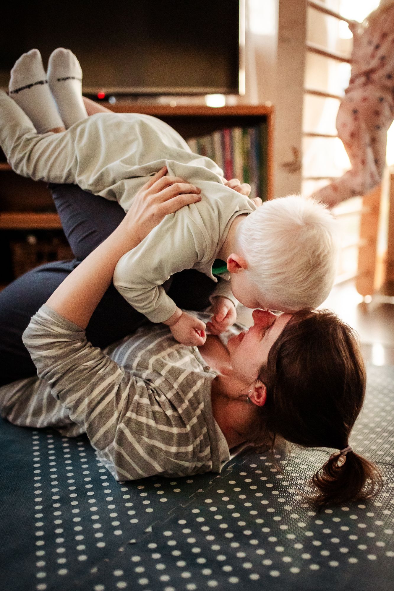 Parent and child share a joyful playful moment lying on a patterned carpet in warm sunlight.