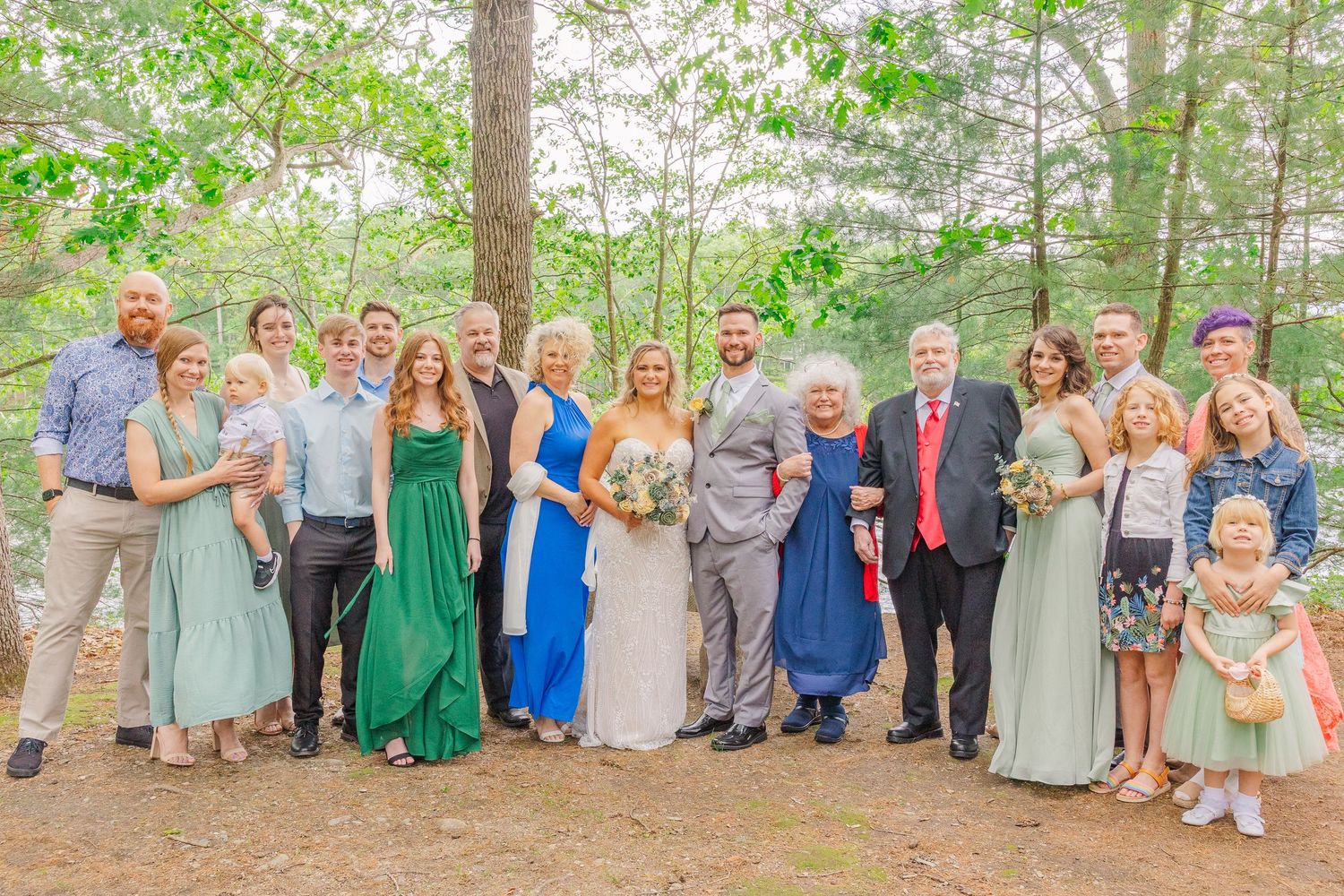 A wedding party in mixed green, blue and neutral colored dresses poses together outdoors in a wooded setting.
