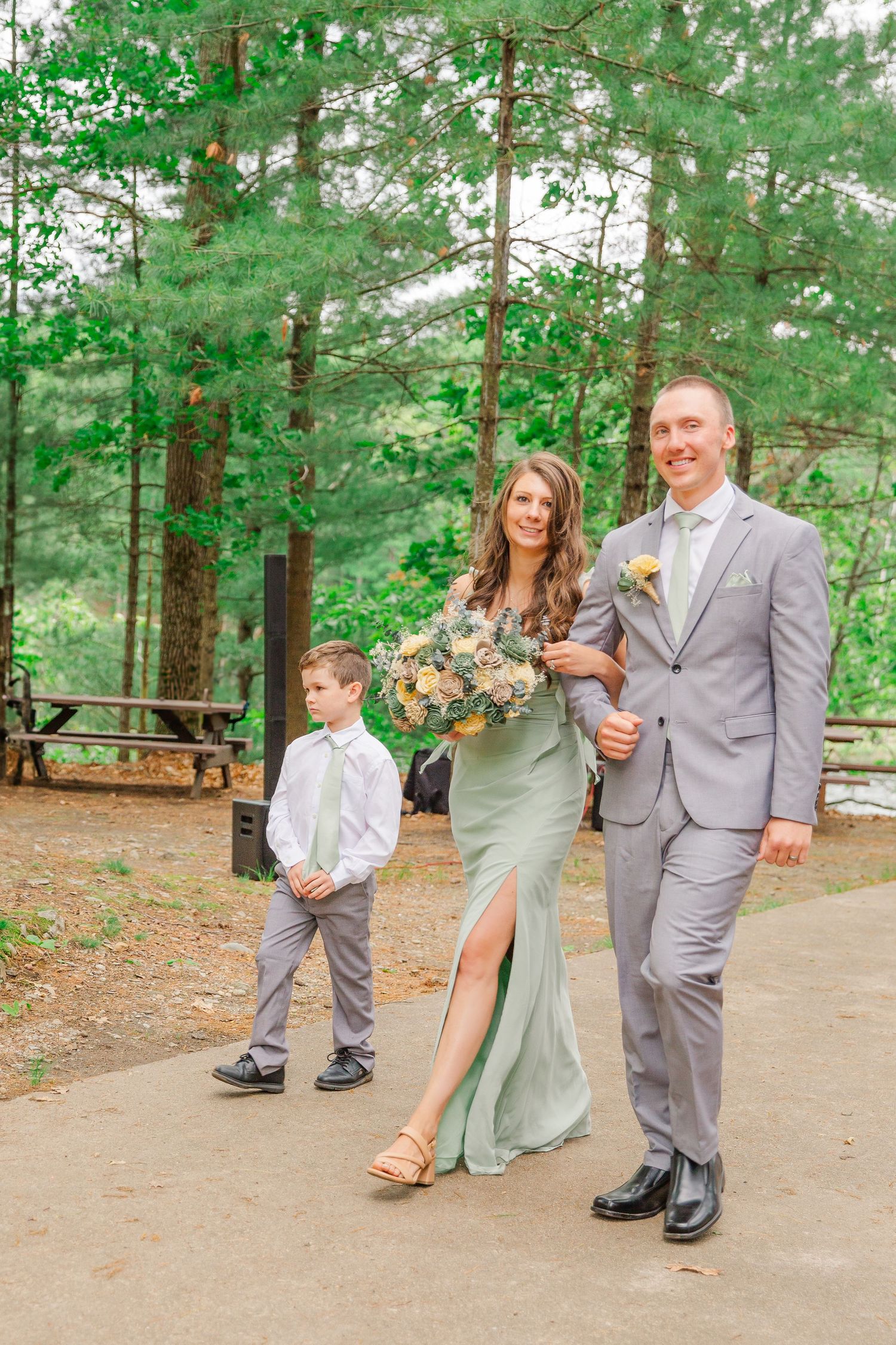 A wedding party in light colored formalwear pose on a path in a pine forest setting holding bouquets.