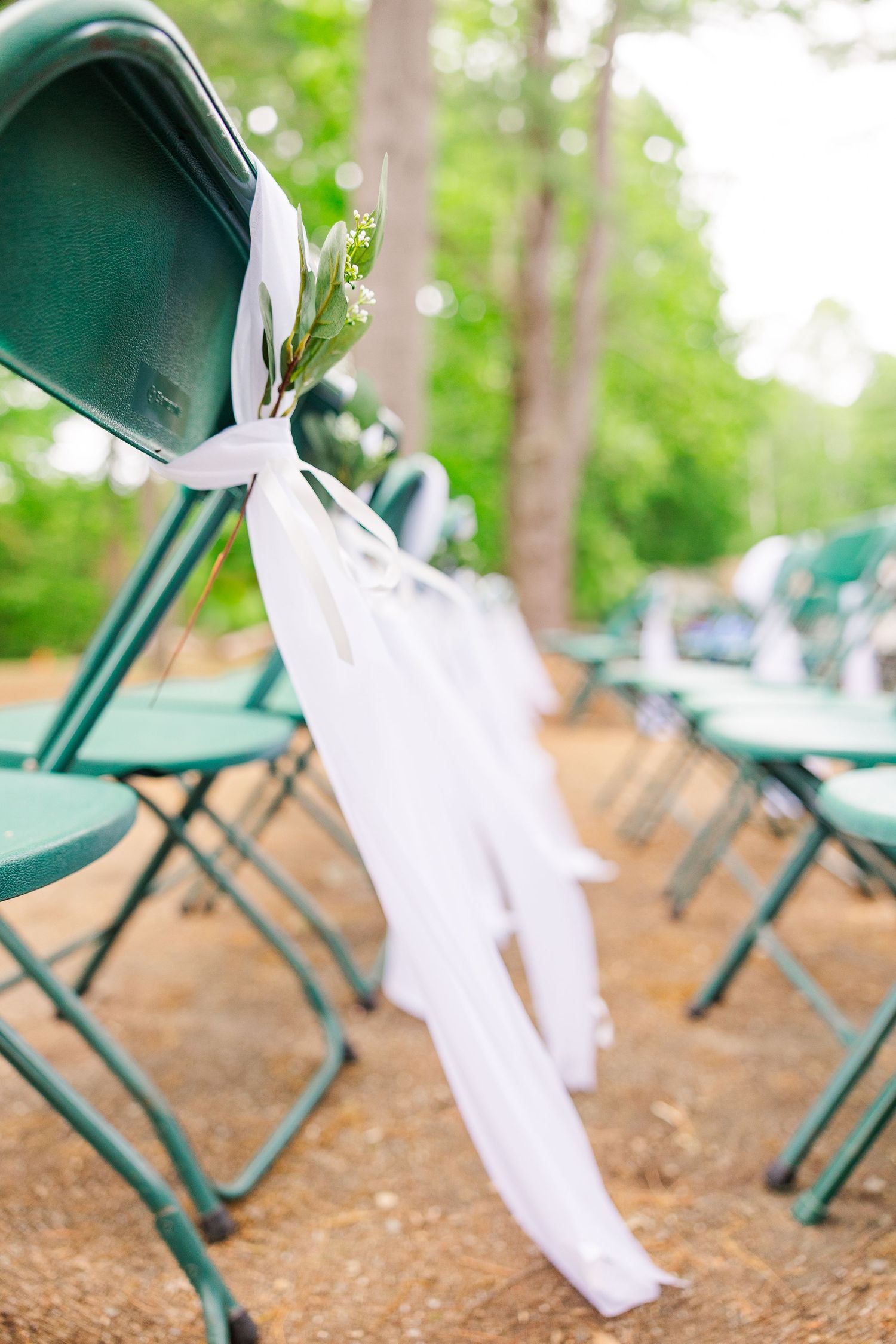 White wedding aisle decor ribbon tied to a green folding chair at an outdoor forest venue.