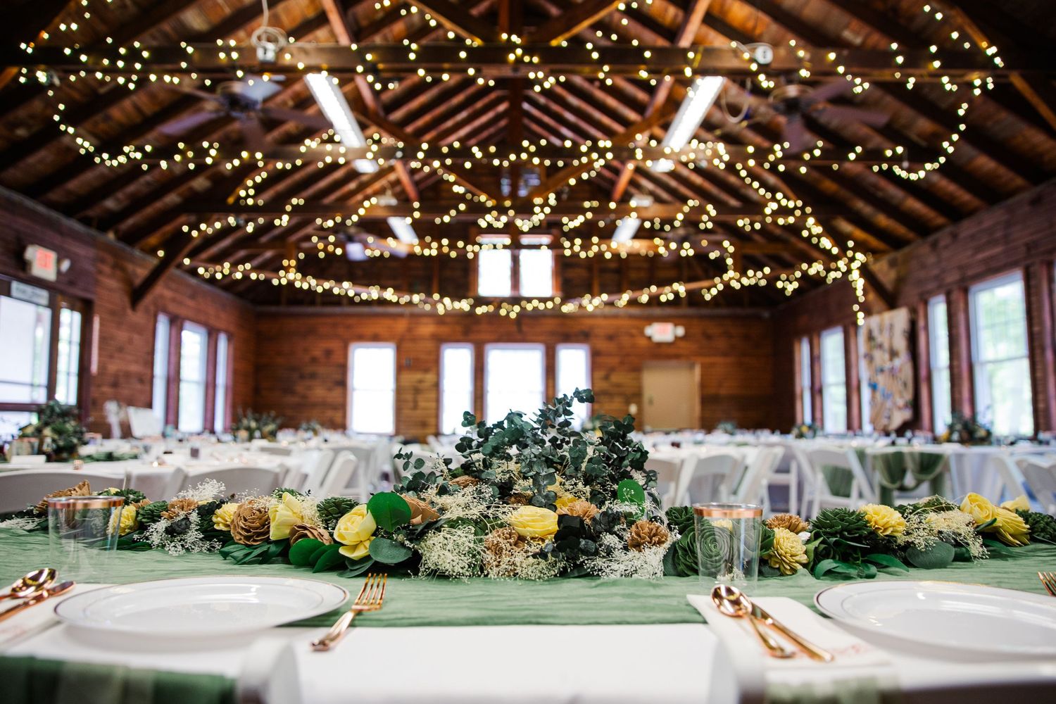 Wedding reception venue decorated with string lights across wooden beams and yellow floral centerpieces on elegant tables.