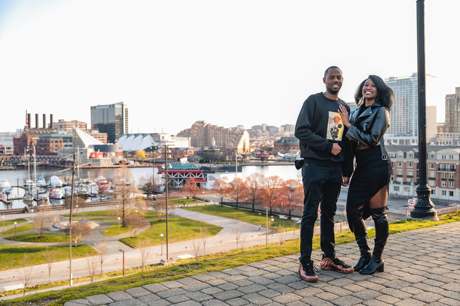 A couple shares a laugh while posing together on a brick path overlooking Baltimore's Inner Harbor on a winter day.