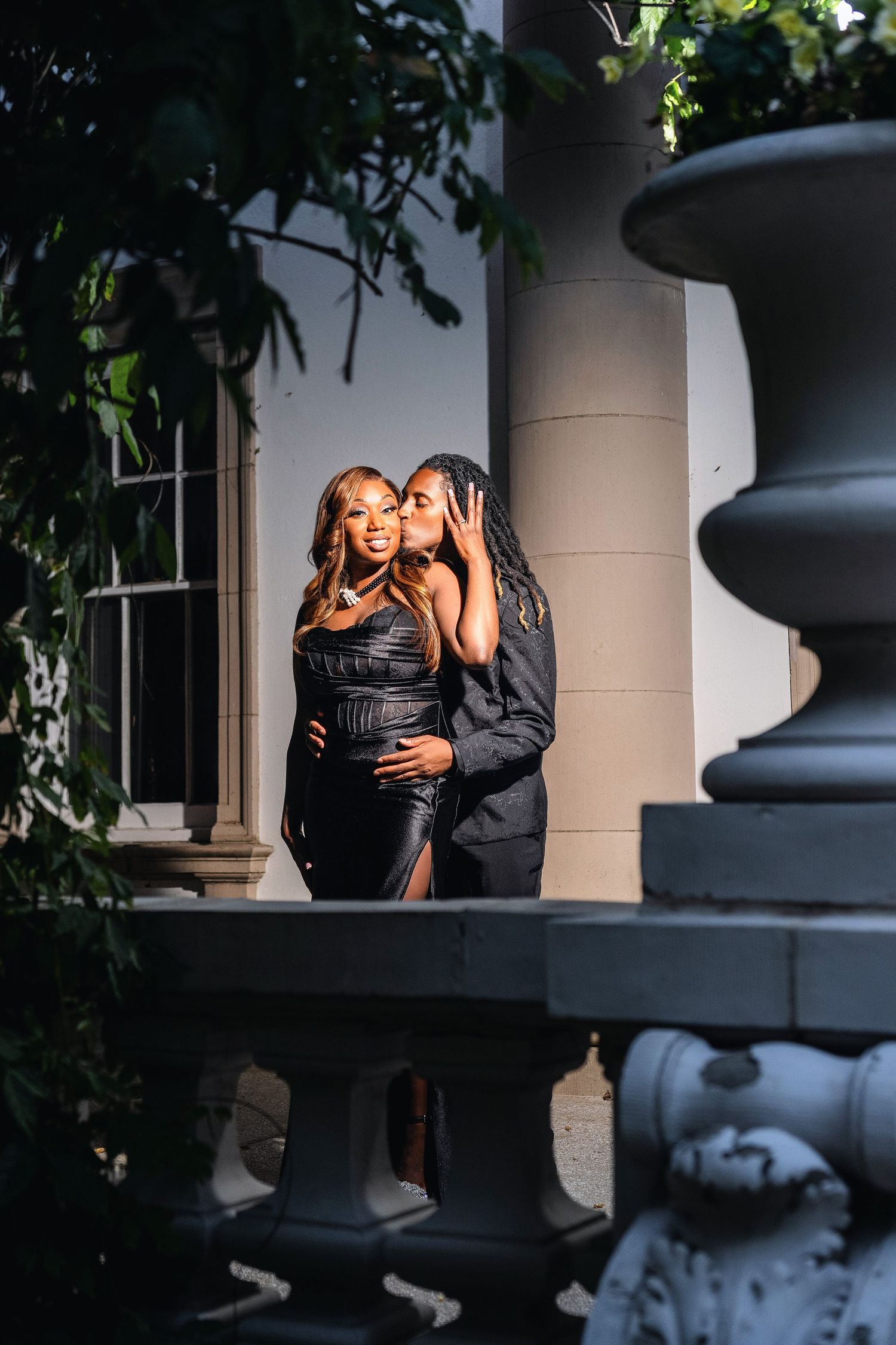 Romantic couple in elegant black attire embrace on an outdoor patio with ornate architectural details at dusk.
