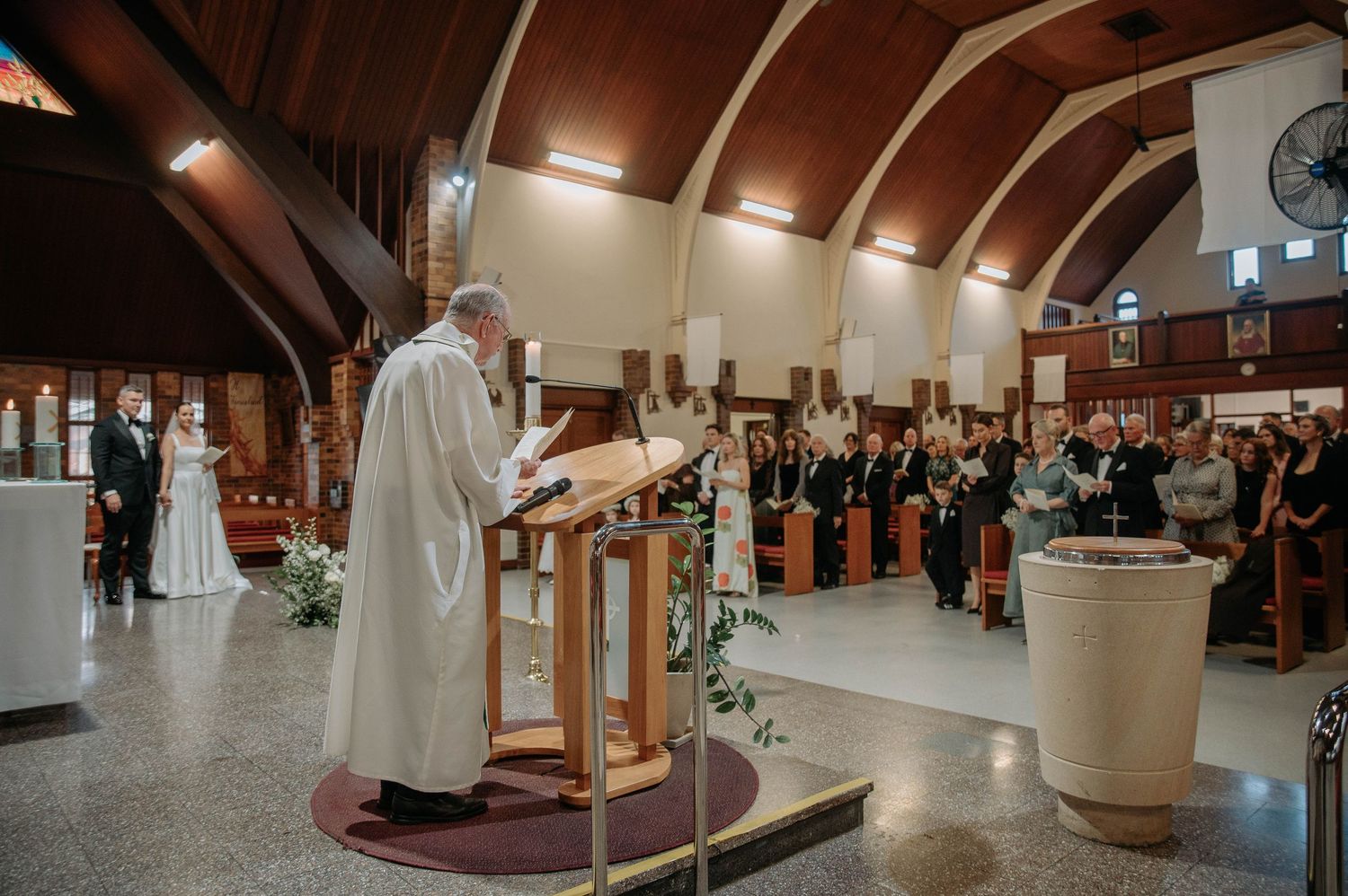 A religious ceremony takes place inside a modern church with vaulted wooden ceilings and rows of standing attendees.