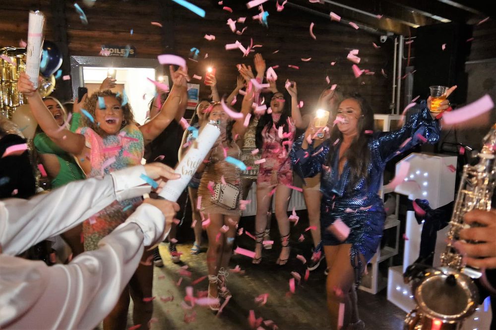 Crowds of party-goers dance and celebrate under falling pink confetti at a festive indoor celebration.