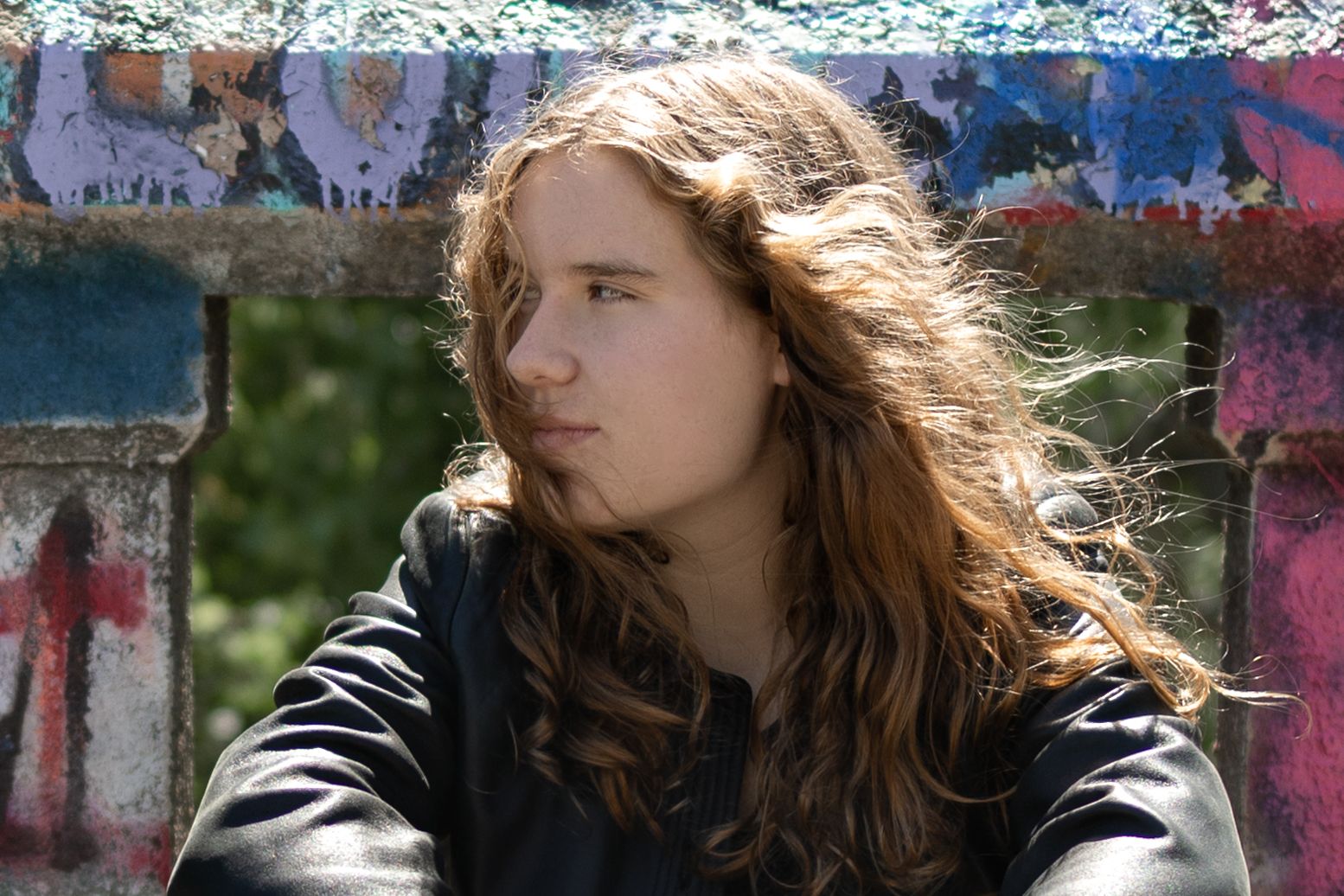 Senior pic of a white female student in black leather jacket and green pants with black boots on a graffiti-covered bridge