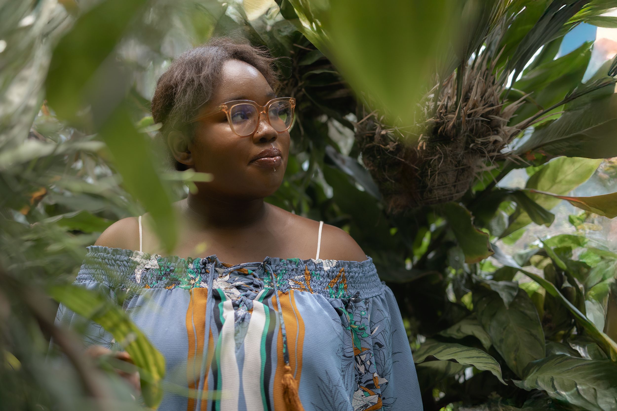 Black female high school student peeking through greenhouse foliage with a striped blue top and trendy glasses