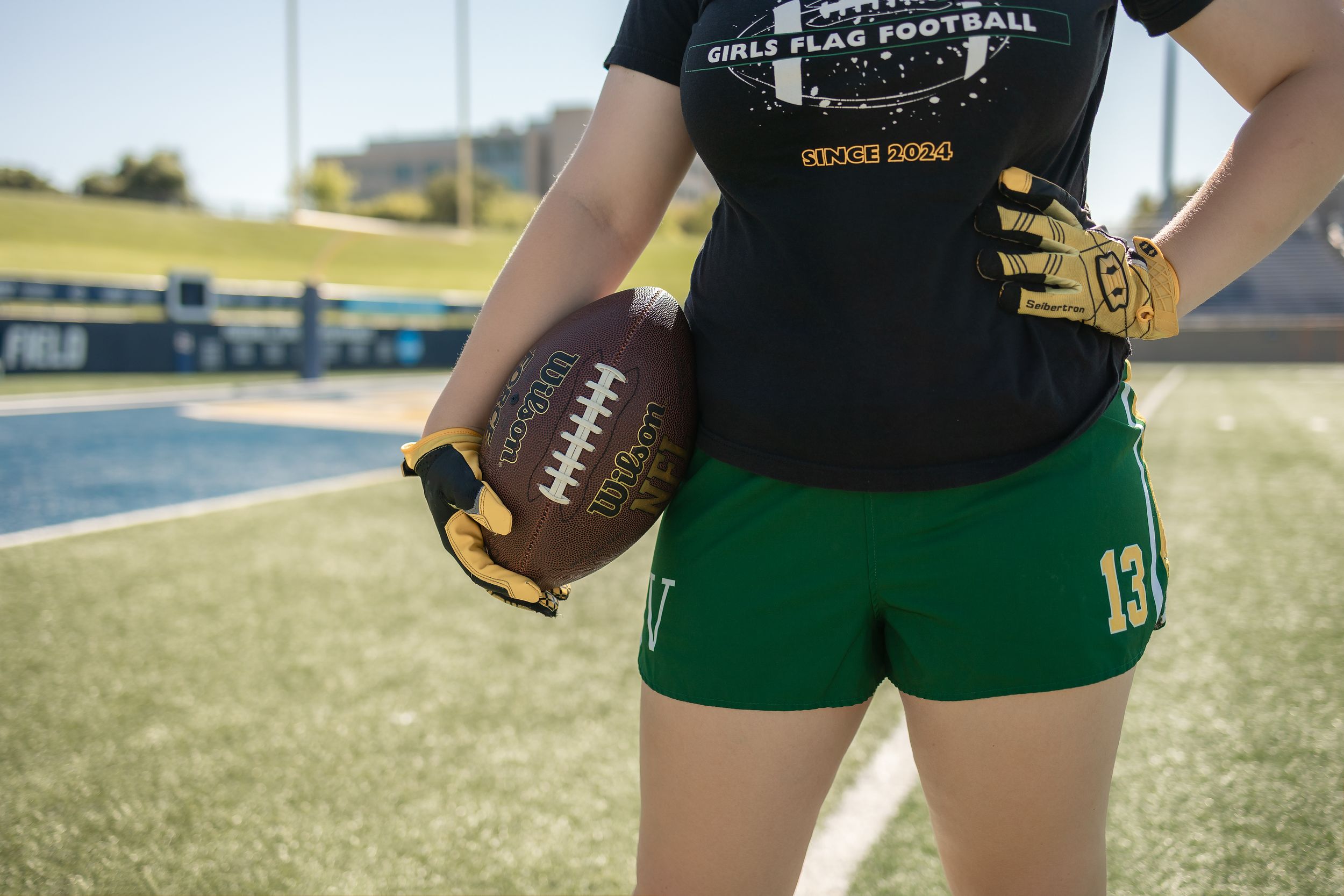 Close-up of female student's midsection to emphasize flag football gloves, uniform, and ball with football field background