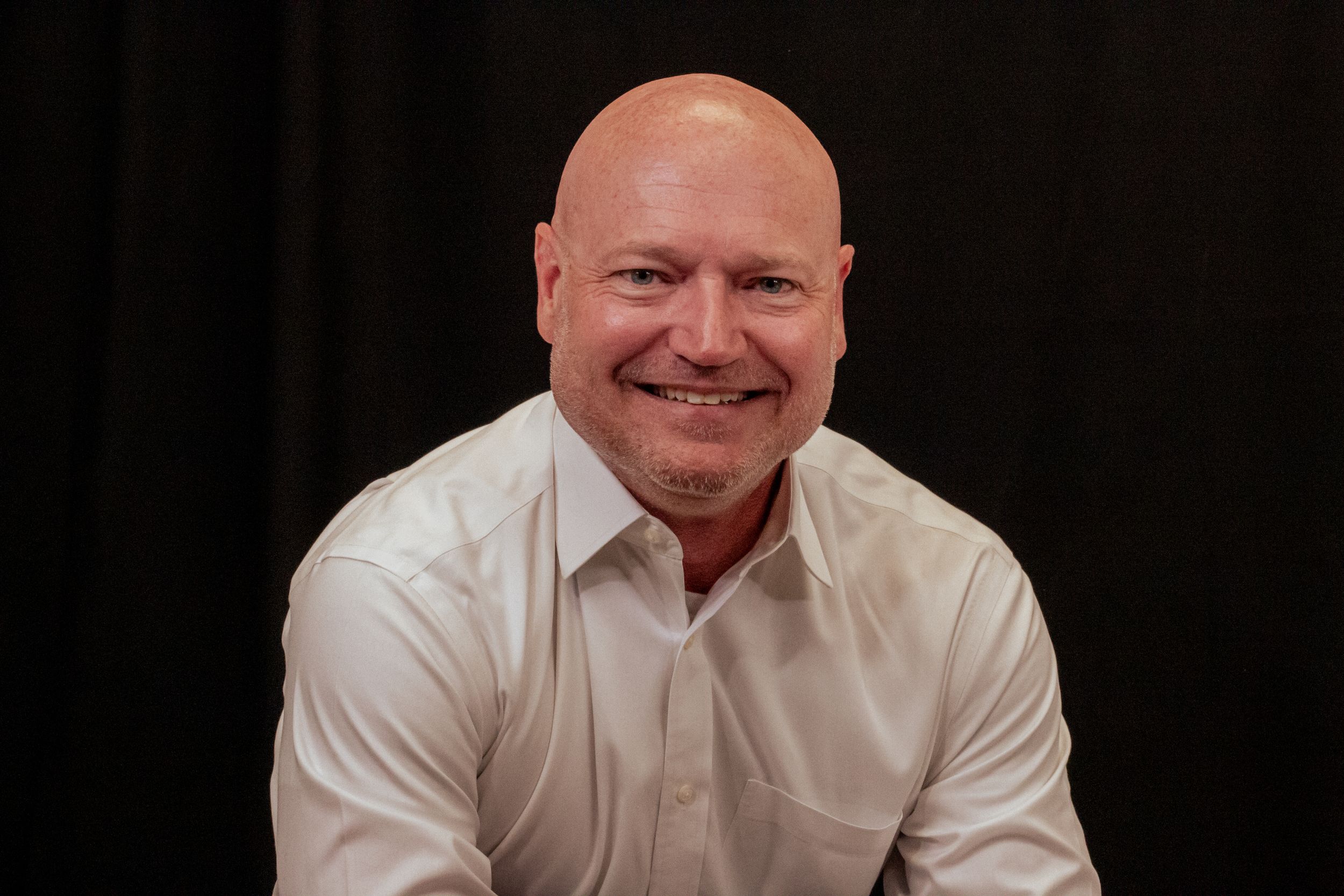A smiling person wearing a white button-down shirt poses for a professional portrait photo against a dark background.