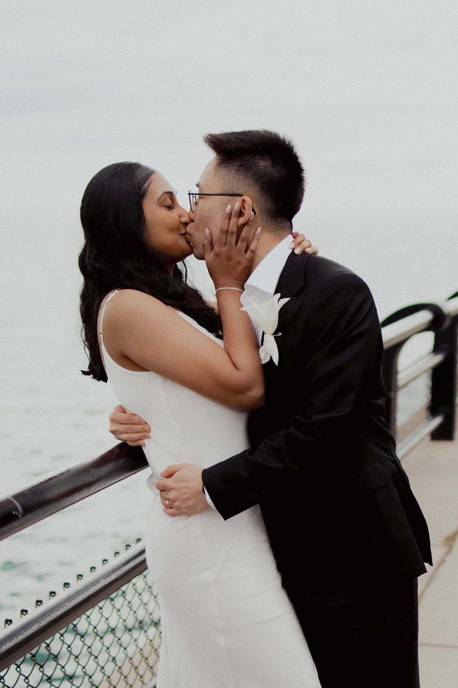 Newlywed couple shares a passionate kiss on a waterfront pier after their wedding ceremony.