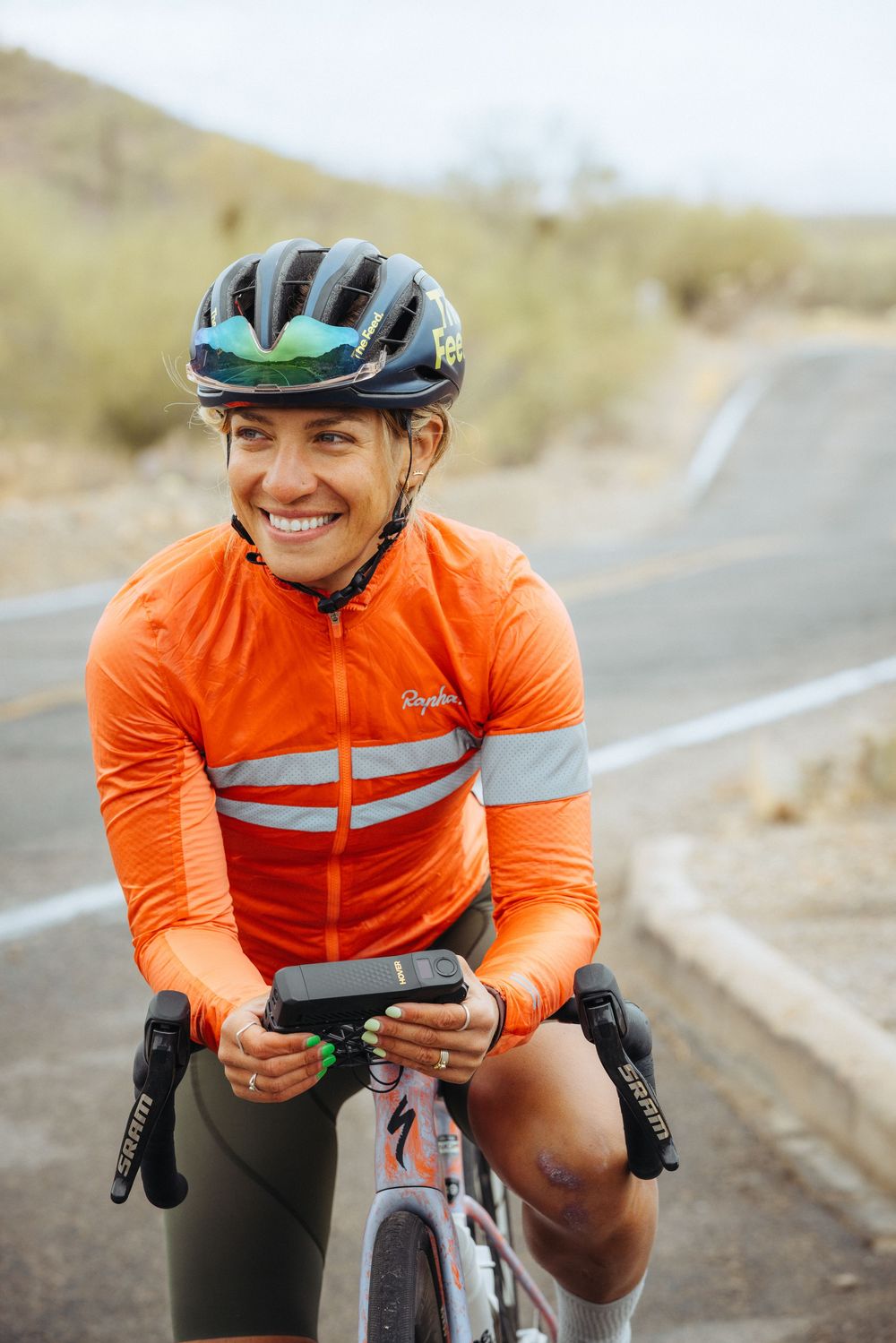 Cyclist in bright orange jacket with reflective stripes on a road bike taking a break on empty desert road.