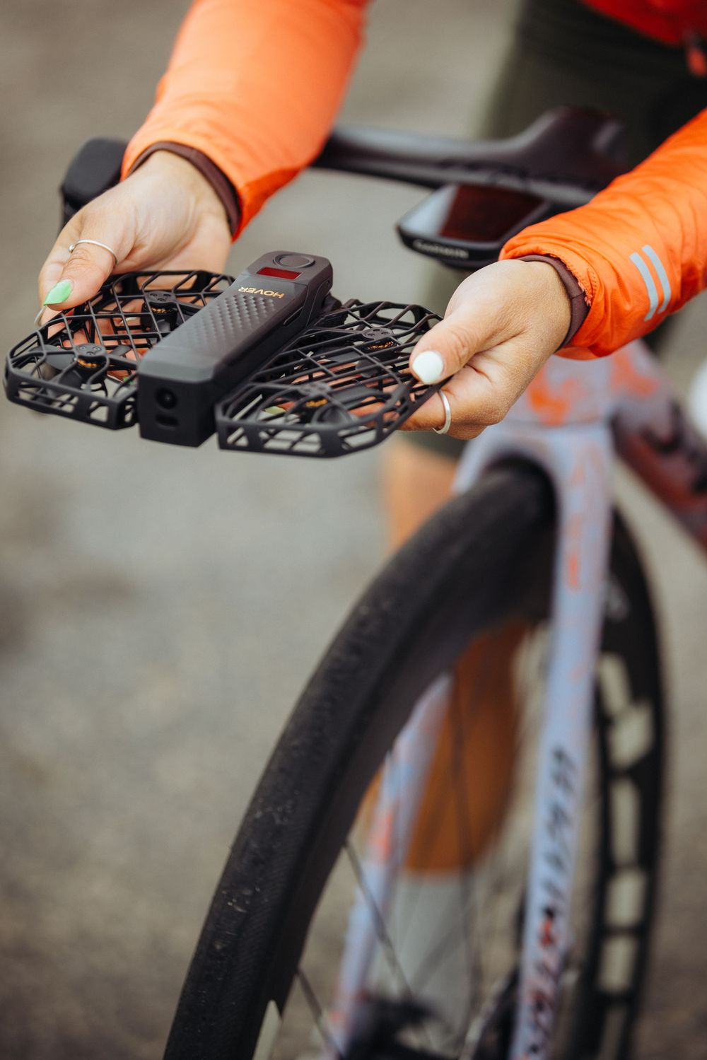 Hands in orange sleeves holding a small black drone or flying device with protective propeller guards.