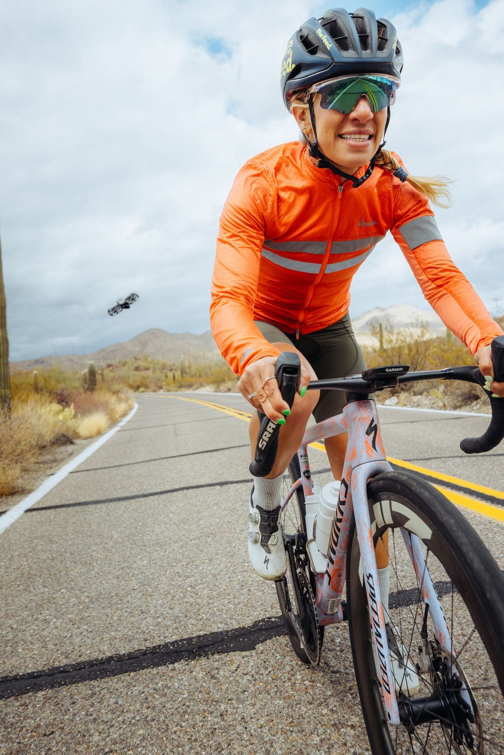Road cyclist in orange jersey riding on a desert road with mountains in the background under cloudy skies.