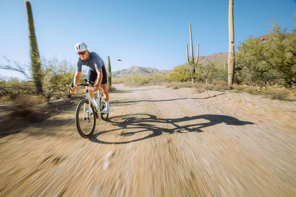 A cyclist rides a road bike on a desert trail surrounded by tall saguaro cacti and mountain views in the background.