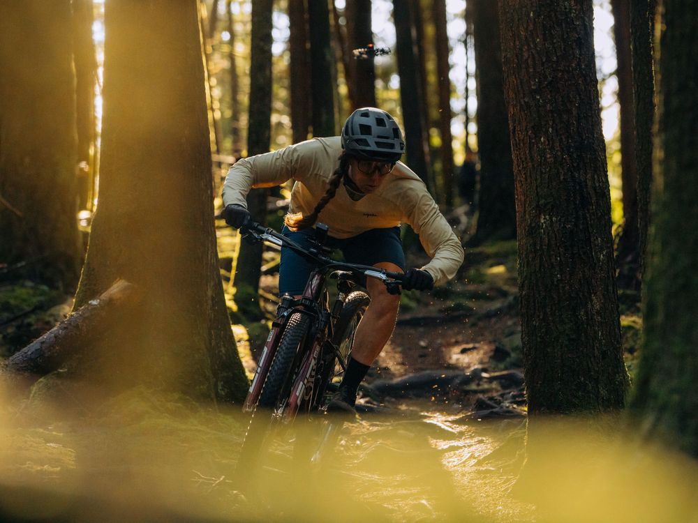 Mountain biker rides through sunlit forest trail during golden hour with trees creating dramatic shadows.