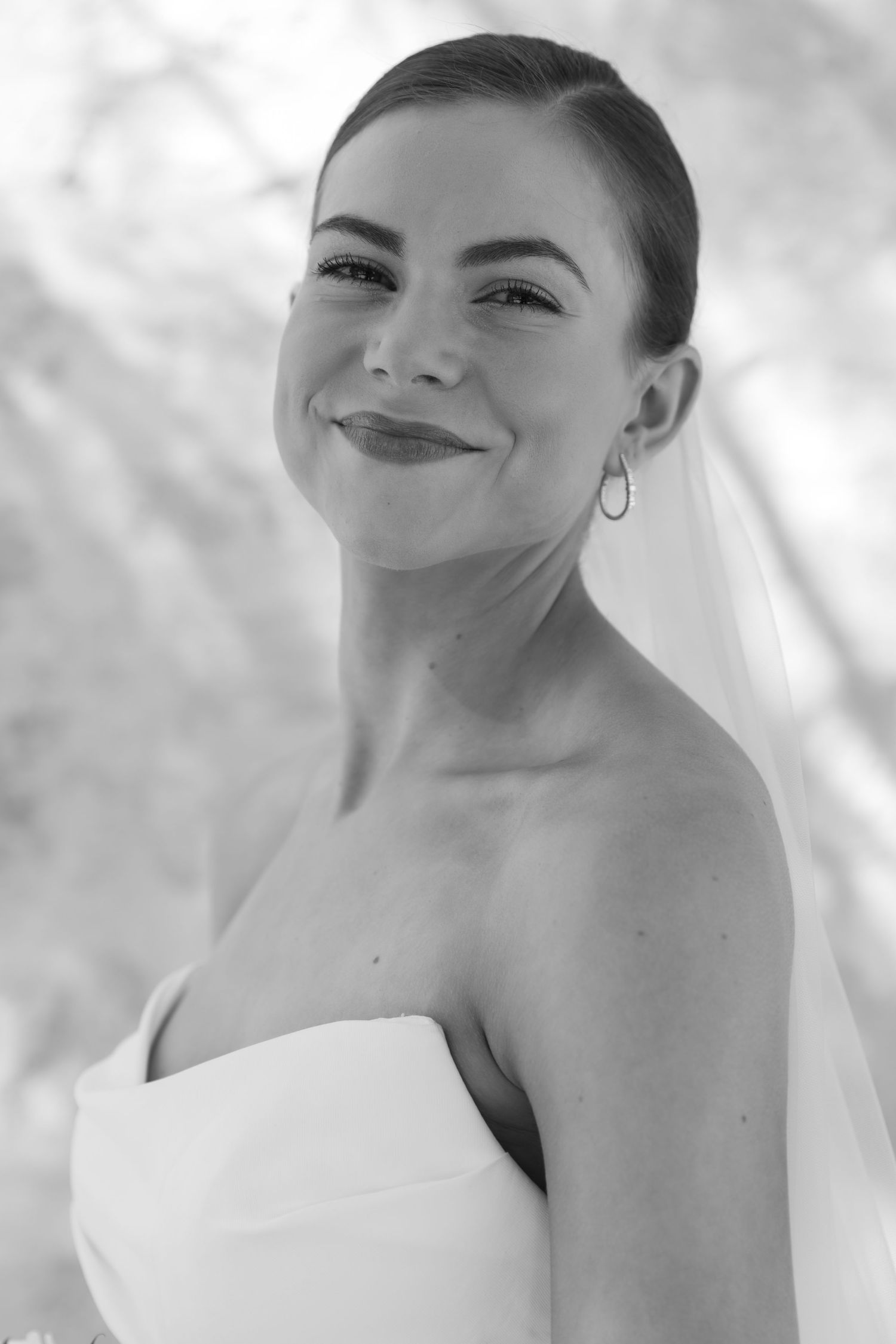 Black and white portrait of a smiling woman in a strapless white dress with sleek updo hairstyle and hoop earrings.