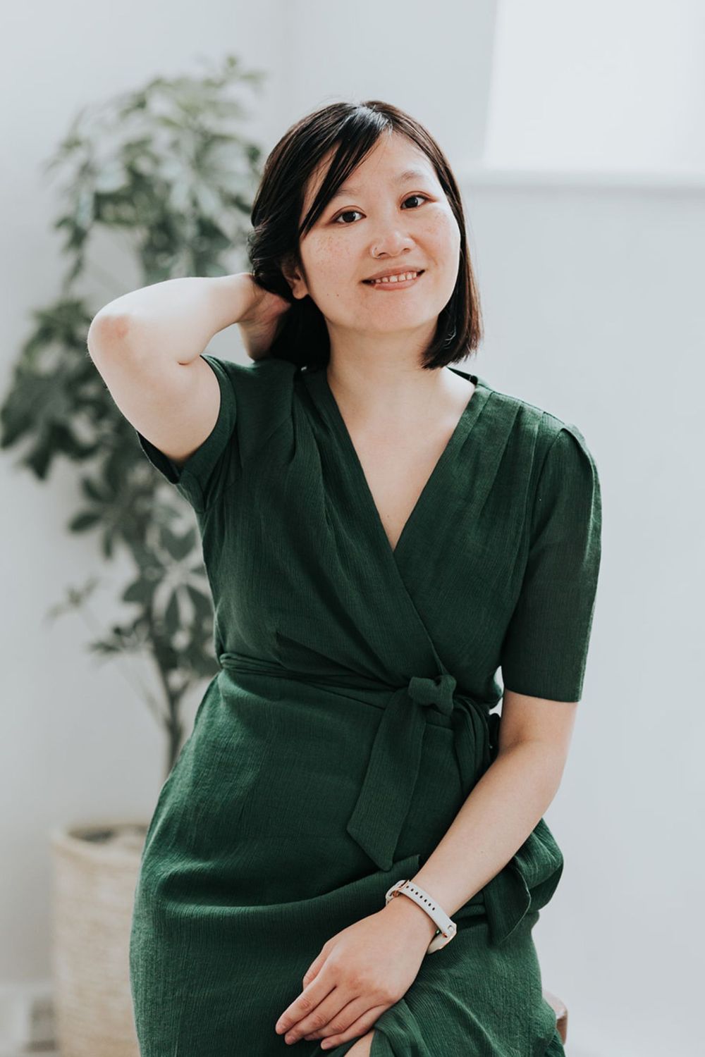 Woman in elegant green wrap dress posing casually against white wall with indoor plant.