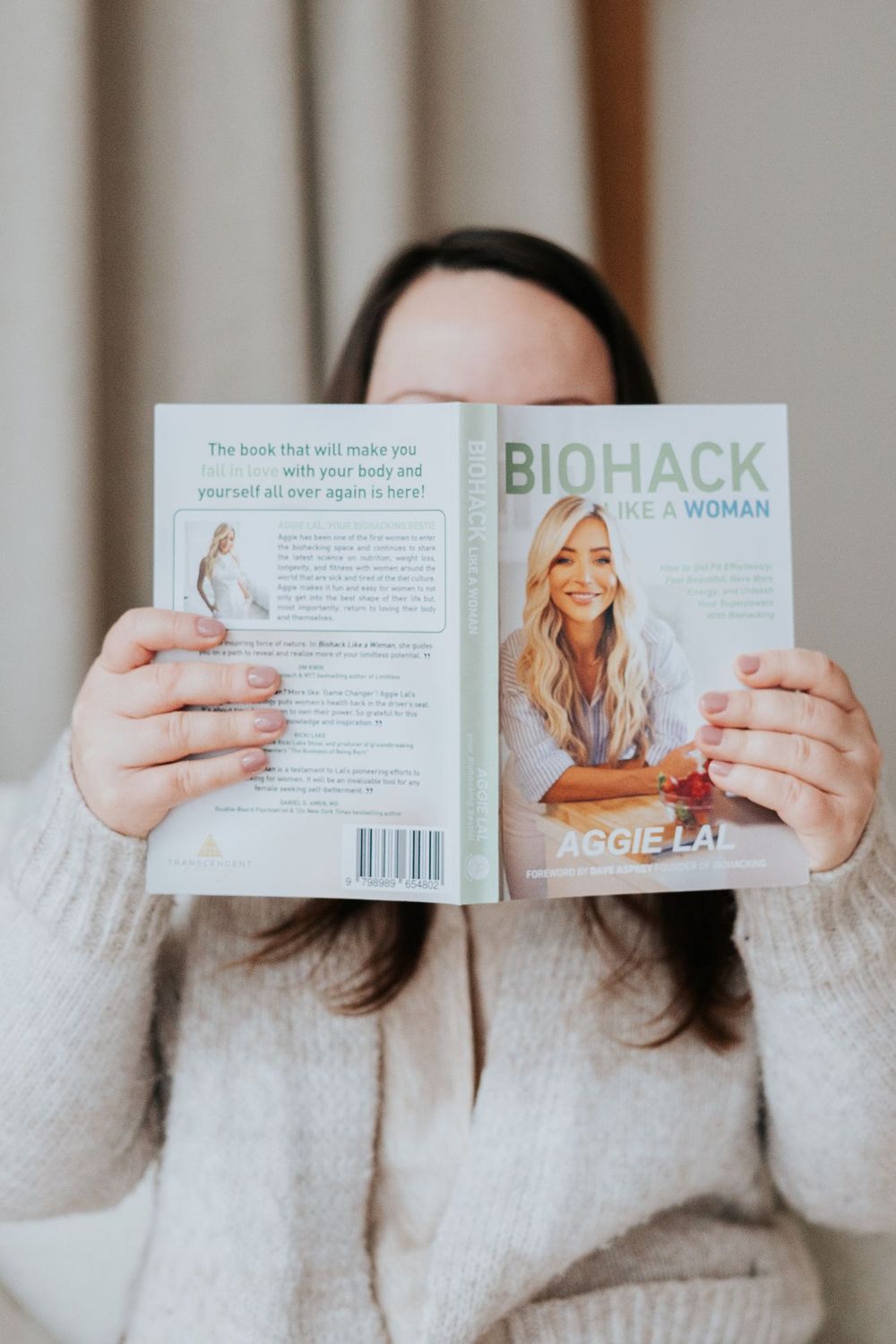 Someone in a cozy cream sweater holds up a wellness book titled 'Biohack for Woman' showing both front and back covers.