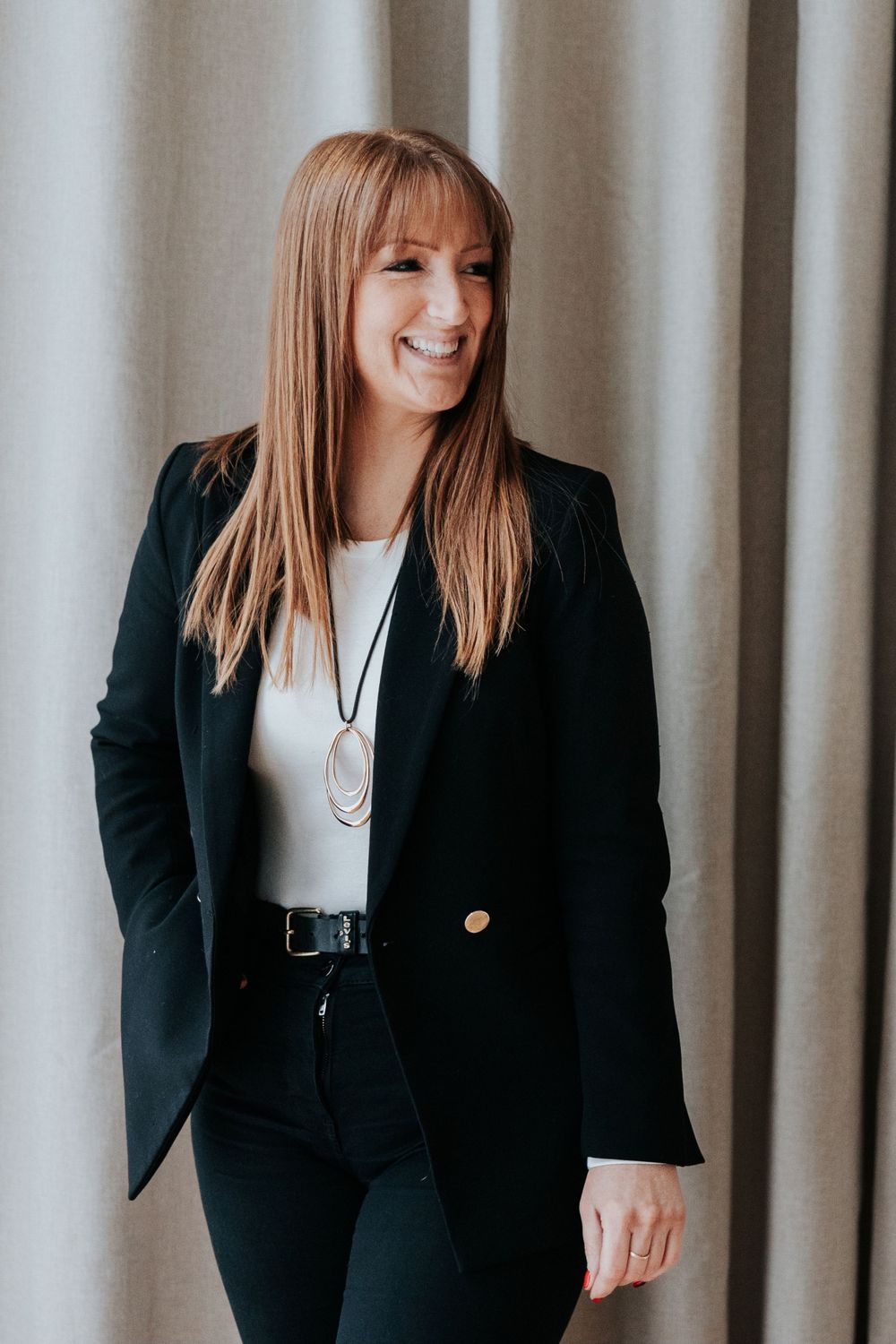 A professional wearing a black blazer and white top smiles warmly while standing against a gray curtain background.