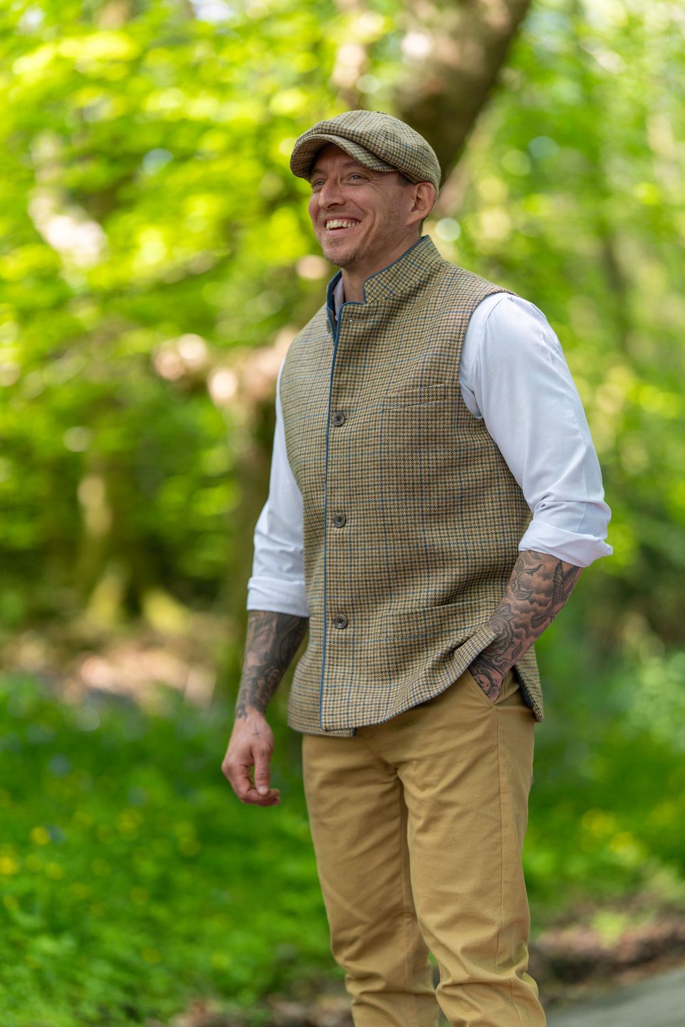 Someone in casual country attire with a tweed vest and khakis stands outdoors in a wooded green setting.