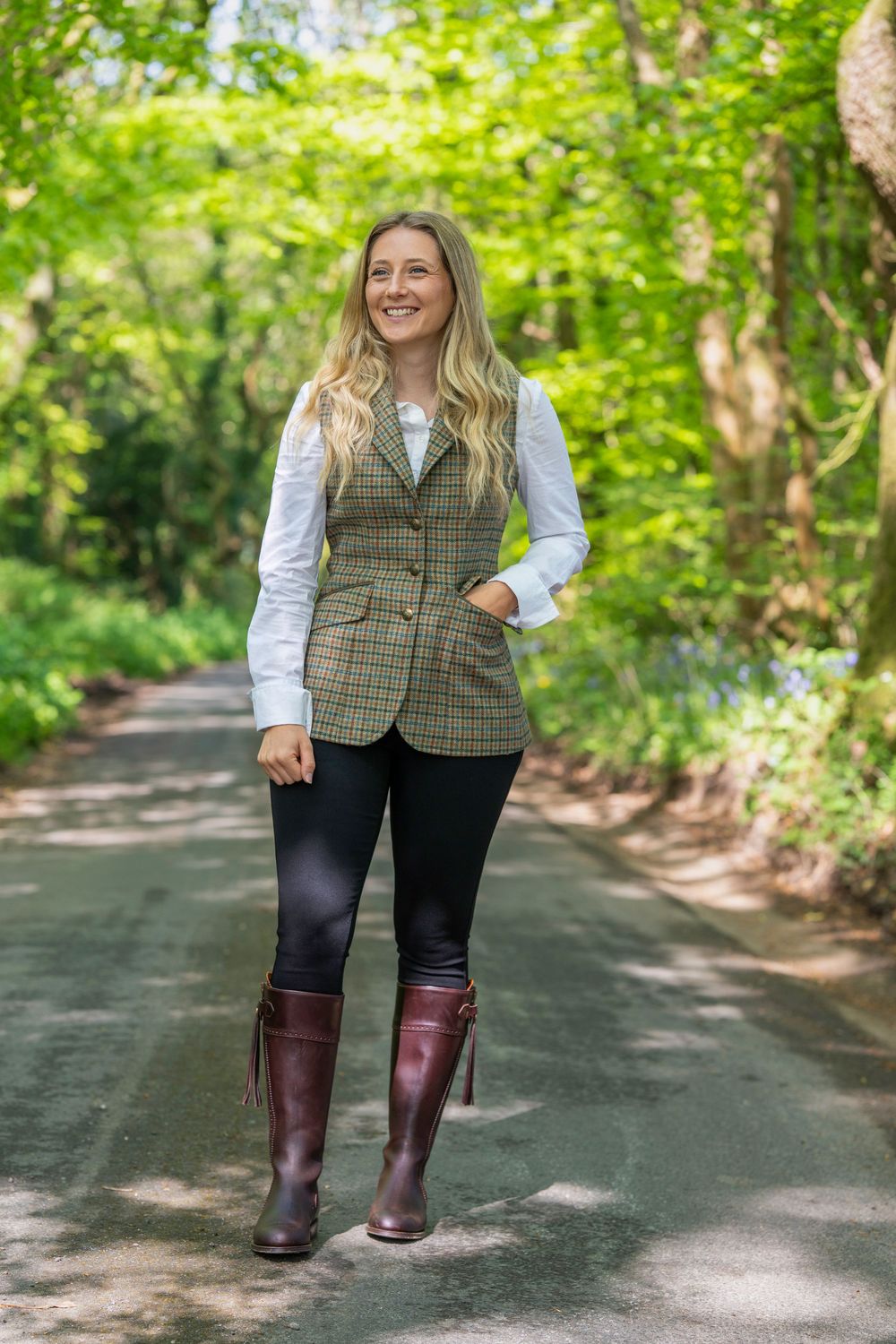 A person wearing a tweed vest, white shirt and burgundy boots walks along a tree-lined path in autumn.